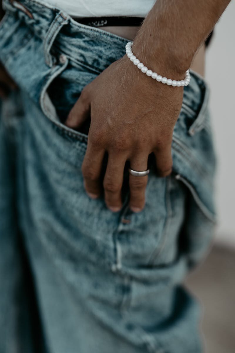 Close-up of a man wearing a pearl bracelet and ring with his hand in a denim pocket.