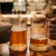 Close-up of two hands clinking beer mugs in a cozy bar atmosphere.