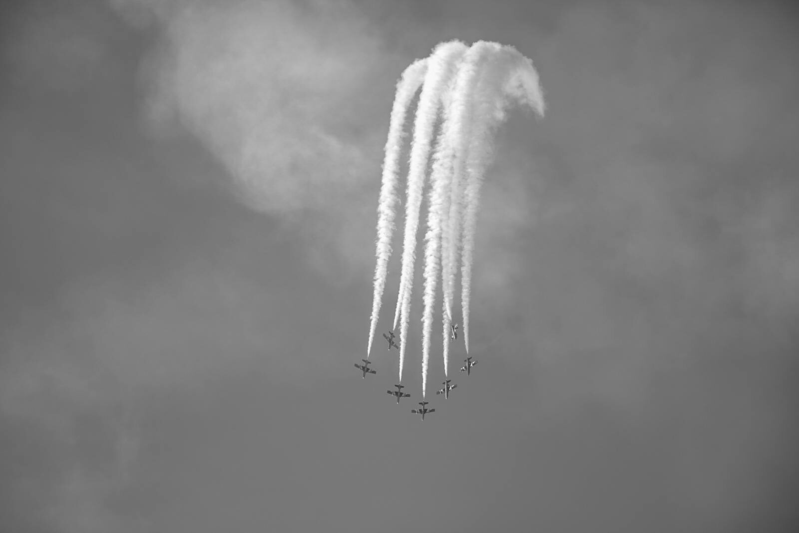Black and white image of an aircraft formation with contrails against a cloudy sky.