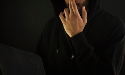 A man in a hoodie and glasses using a laptop in a dark room, representing cyber security.