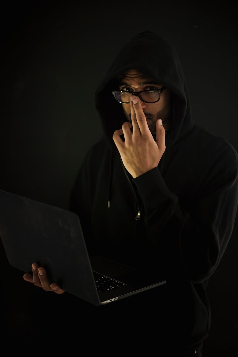 A man in a hoodie and glasses using a laptop in a dark room, representing cyber security.