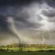A powerful tornado and vibrant lightning striking over a rural countryside landscape.