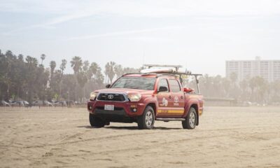 A red truck parked on the sandy beach of Santa Monica, California under a bright sky.