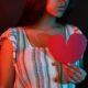 Young woman holding a red paper heart, portrayed in dramatic red and blue lighting.