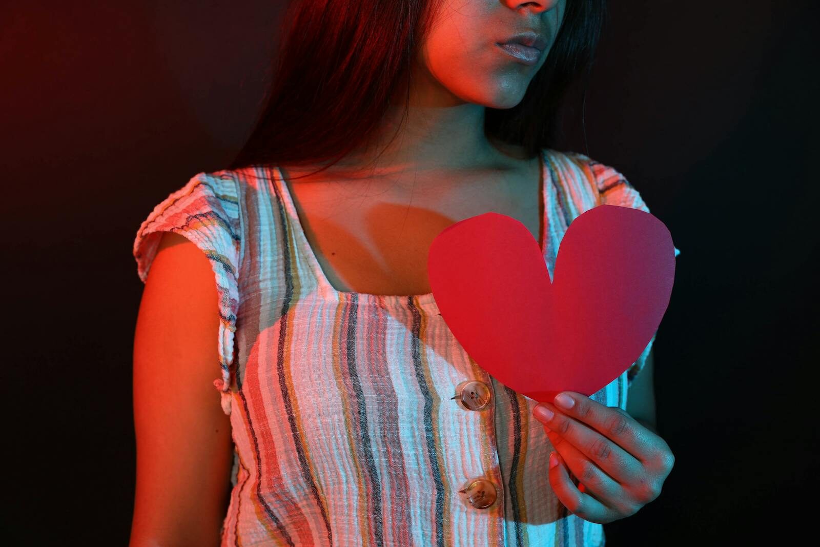 Young woman holding a red paper heart, portrayed in dramatic red and blue lighting.