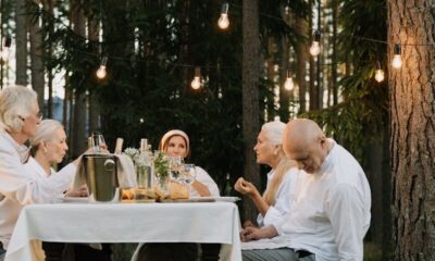 Senior adults enjoying an outdoor meal with a pet corgi under string lights in a forest setting.