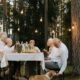 Senior adults enjoying an outdoor meal with a pet corgi under string lights in a forest setting.