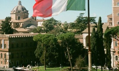 Italian flag waving over Rome cityscape with historic buildings and pedestrians in summer.