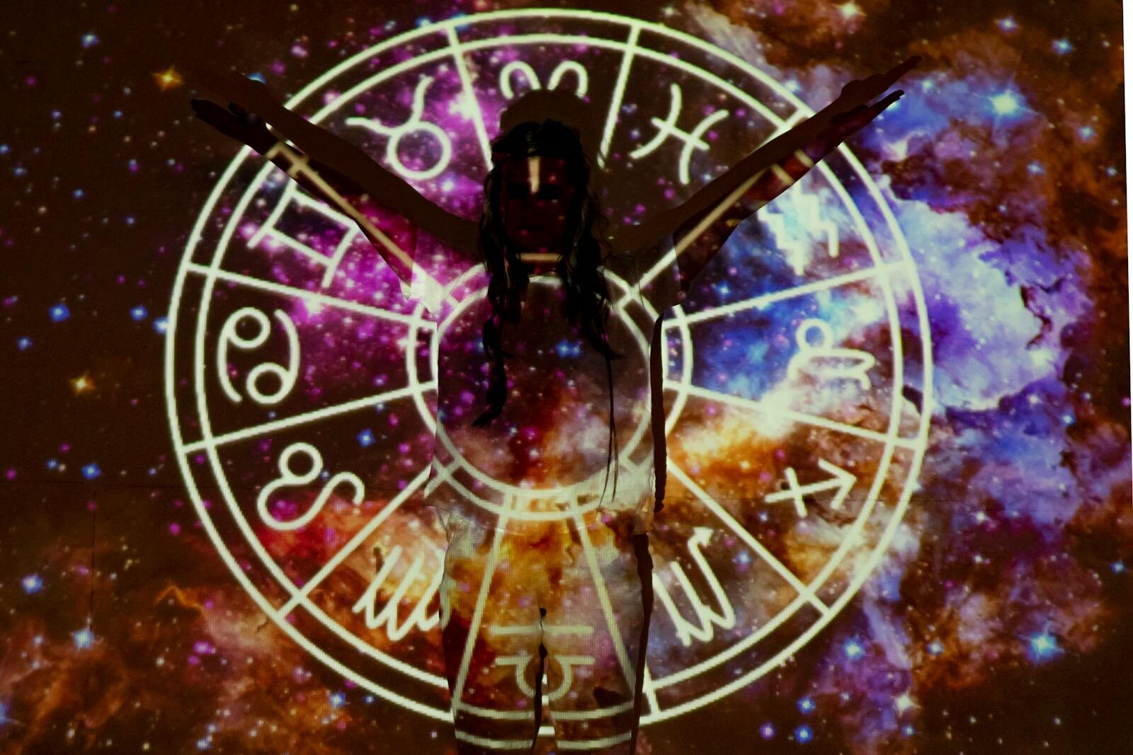 Silhouette of person extending arms against zodiac symbols and cosmic background, esoteric theme.
