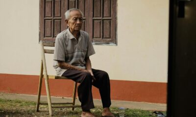 Elderly man sitting pensively on a wooden chair outside his home, reflecting solitude.