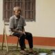Elderly man sitting pensively on a wooden chair outside his home, reflecting solitude.