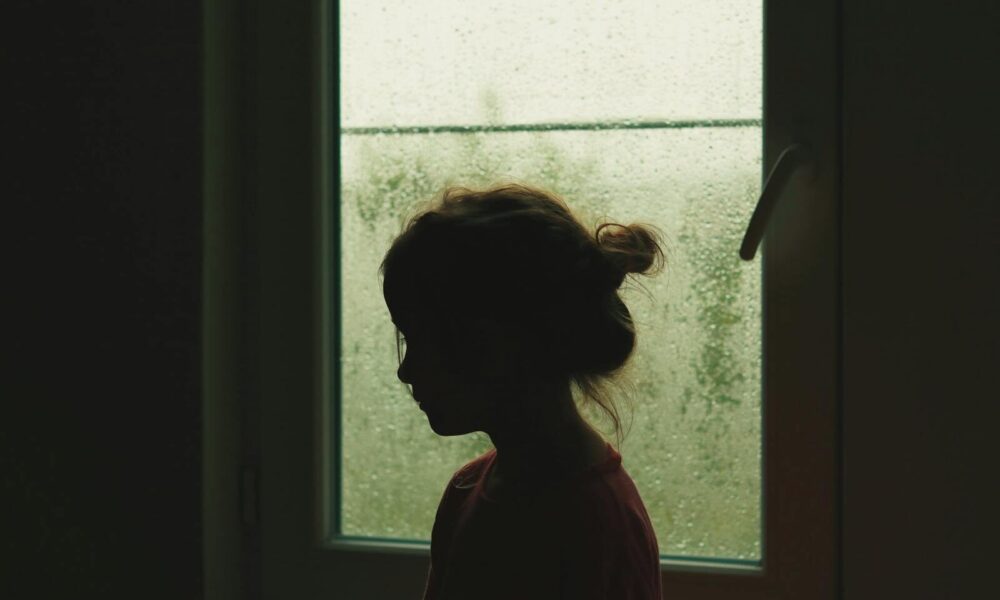 A child's silhouette stands by a rainy window, creating a nostalgic indoor scene.