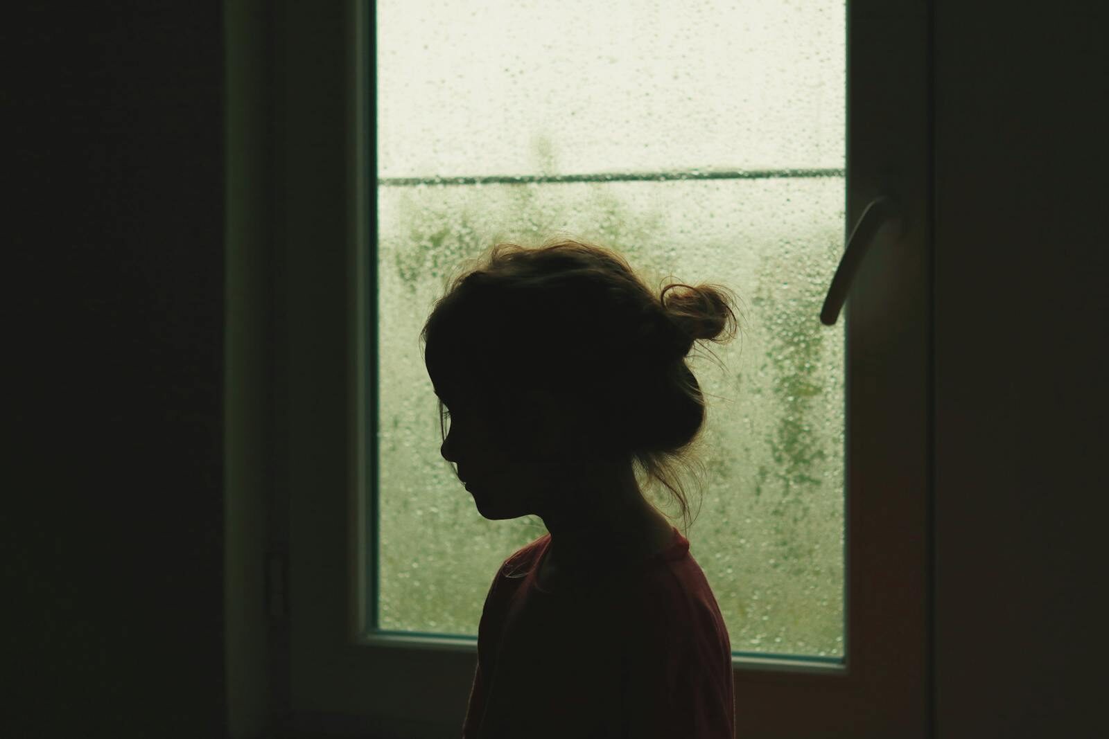 A child's silhouette stands by a rainy window, creating a nostalgic indoor scene.