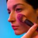 Close-up of a woman applying makeup with a brush against a vibrant backdrop.