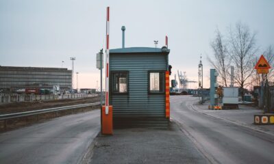 Industrial checkpoint and barrier at port under cloudy sky with cranes in the background.