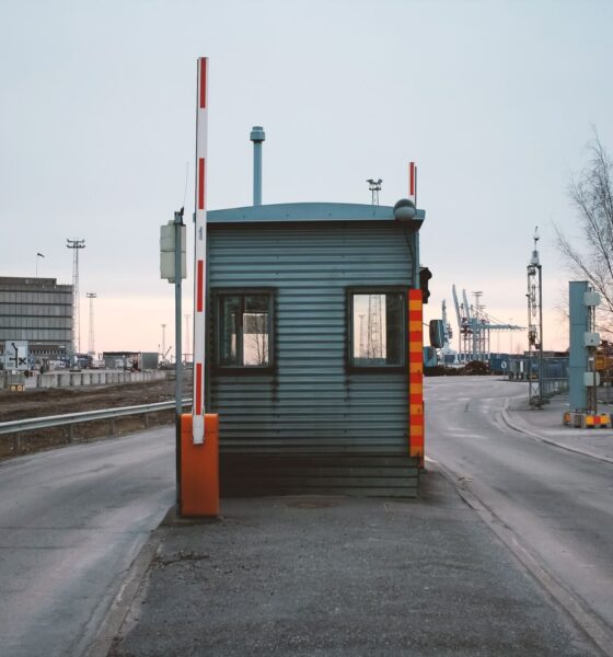 Industrial checkpoint and barrier at port under cloudy sky with cranes in the background.