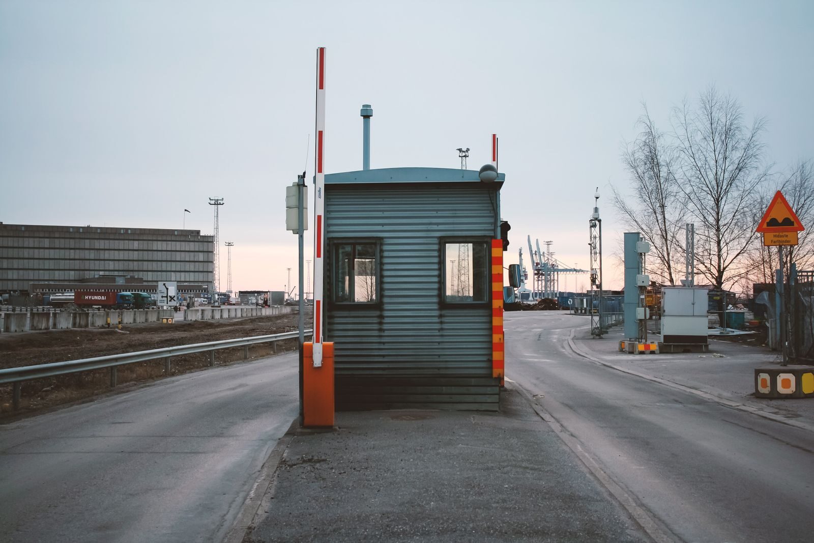 Industrial checkpoint and barrier at port under cloudy sky with cranes in the background.