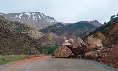 A scenic mountain road blocked by large rocks from a landslide with a snowy peak in the background.