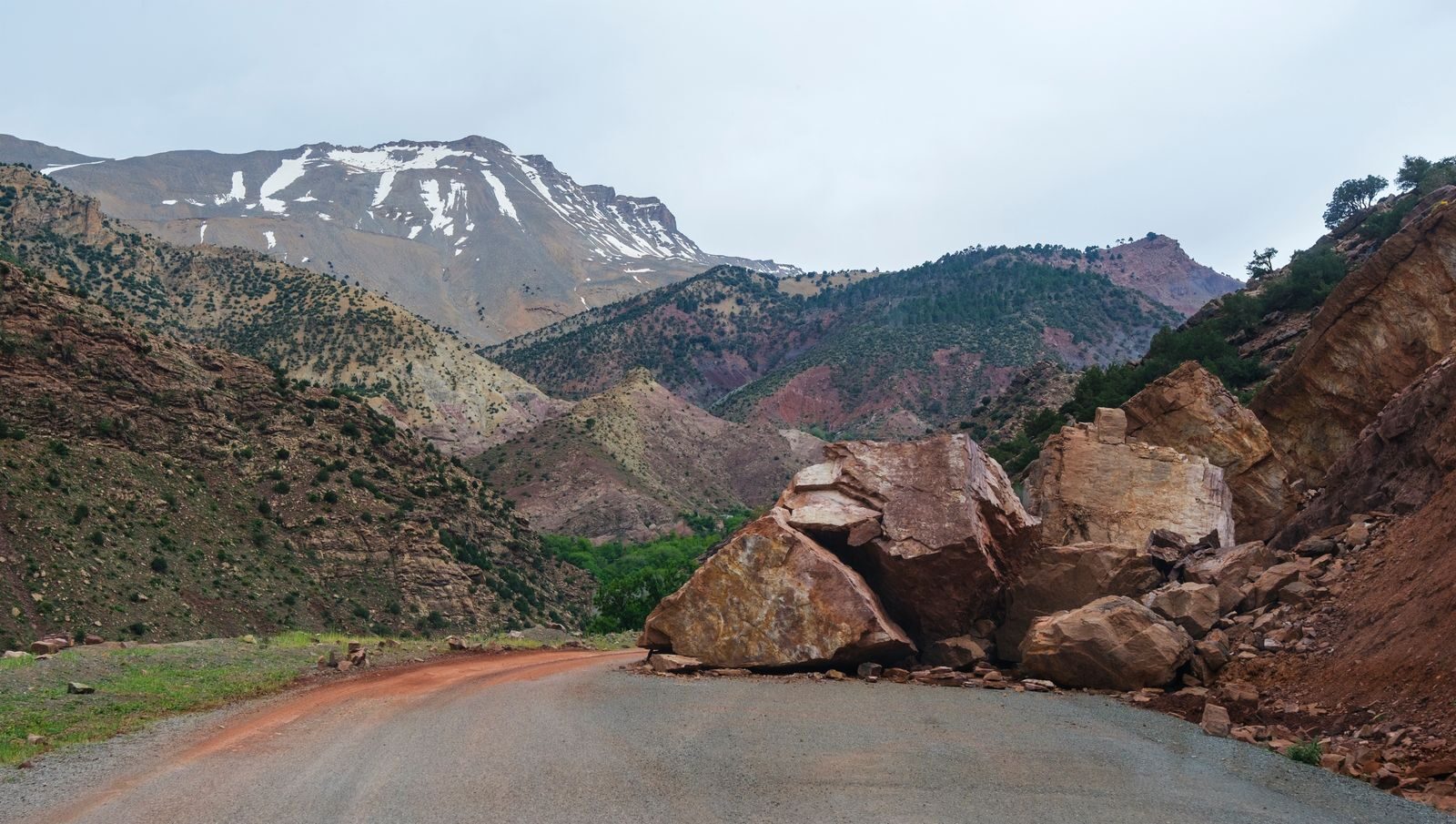 A scenic mountain road blocked by large rocks from a landslide with a snowy peak in the background.