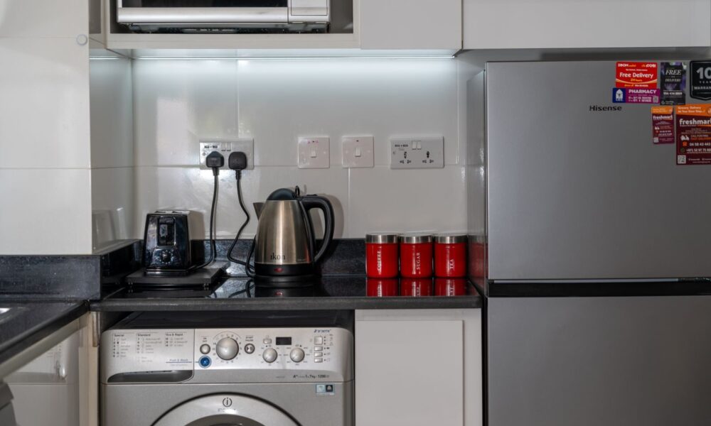 Stylish modern kitchen with stainless steel appliances and red accents, highlighting a washing machine, fridge, and countertop electronics.