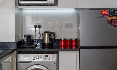 Stylish modern kitchen with stainless steel appliances and red accents, highlighting a washing machine, fridge, and countertop electronics.