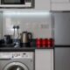 Stylish modern kitchen with stainless steel appliances and red accents, highlighting a washing machine, fridge, and countertop electronics.