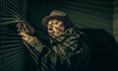 A man in a hat suspiciously peering through blinds, holding a phone in shadowy light.