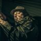 A man in a hat suspiciously peering through blinds, holding a phone in shadowy light.