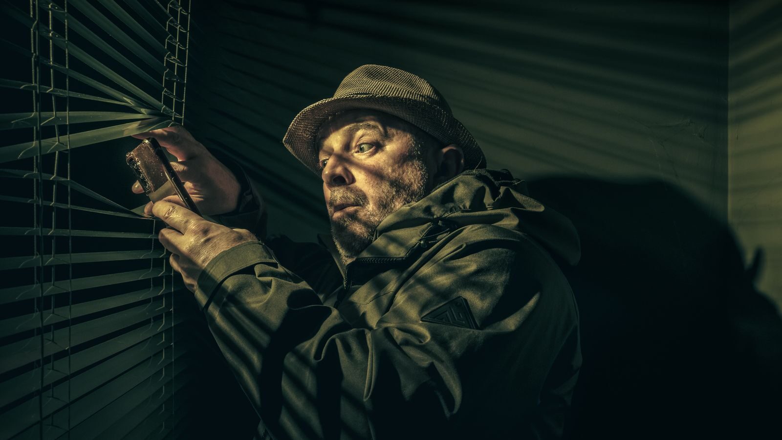 A man in a hat suspiciously peering through blinds, holding a phone in shadowy light.