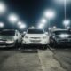 Night scene in a well-lit parking lot featuring various parked cars and street lamps.