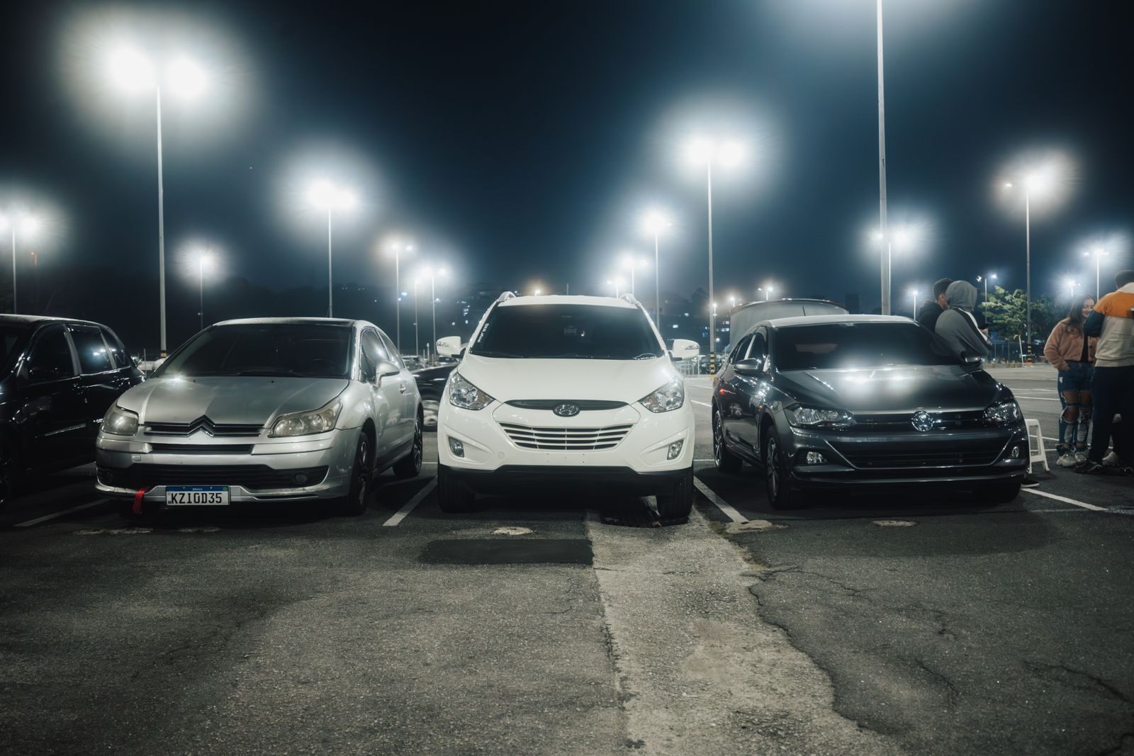 Night scene in a well-lit parking lot featuring various parked cars and street lamps.