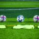 Three soccer balls and training bibs on a lush green field in Rawalpindi, Pakistan.