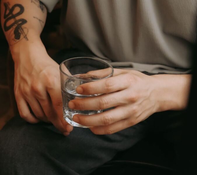 A seated man with tattooed arm holding a glass of water, indoors setting.