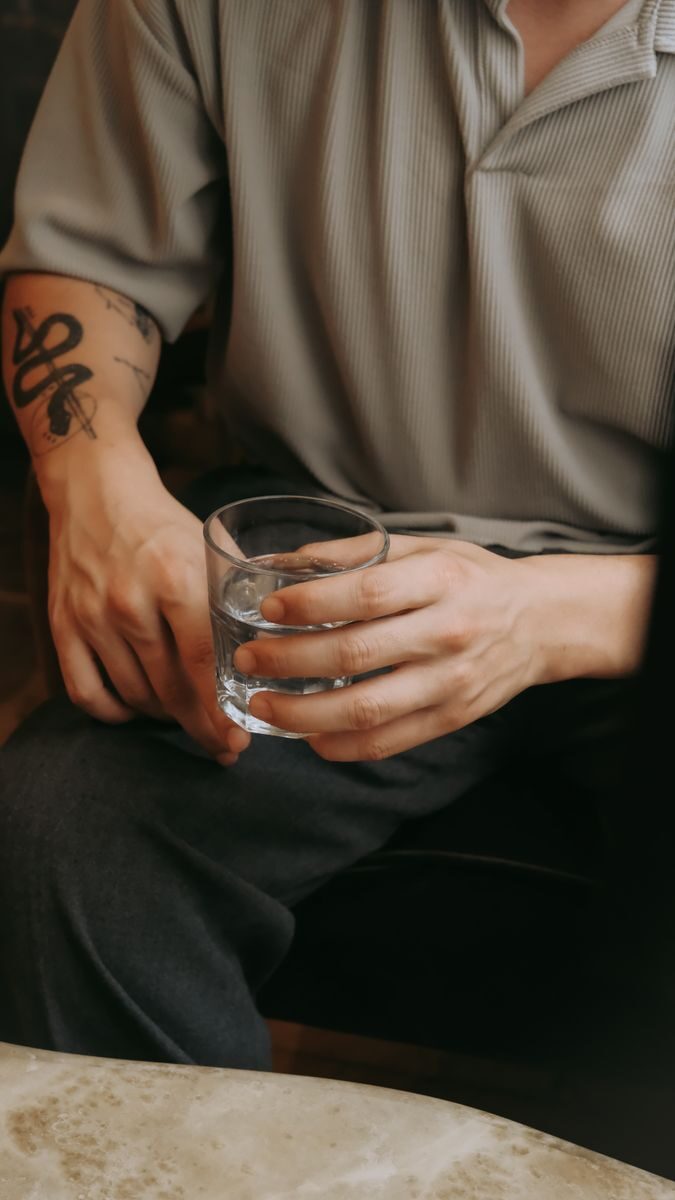 A seated man with tattooed arm holding a glass of water, indoors setting.