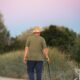 Man in green shirt and hat walking on a rural path with a stick at sunset.
