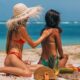 Two women enjoying sunbathing on a tropical beach, applying lotion while relaxing in swimwear.