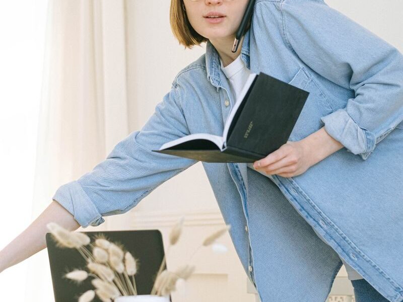 A young woman multitasks with a phone call and notebook in a casual home office setting.