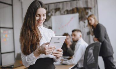 Young woman in a professional office setting browsing a tablet with colleagues in the background.