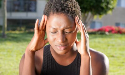 A woman holding her head in pain, indicating a severe headache outdoors in a sunny setting.