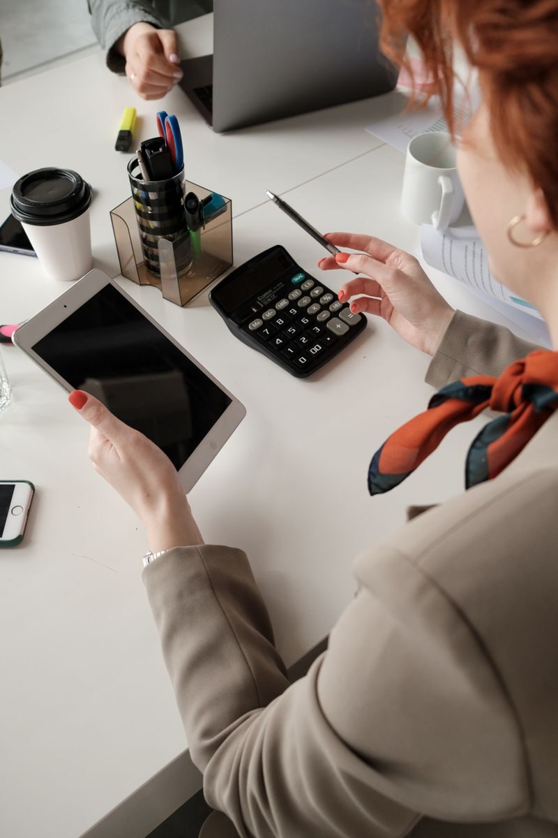 A businesswoman in an office setting holds a tablet while calculating with a pen and calculator on the table.