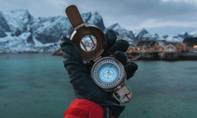 A gloved hand holds a compass in Reine, Lofoten Islands, with a stunning fjord backdrop.