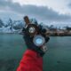 A gloved hand holds a compass in Reine, Lofoten Islands, with a stunning fjord backdrop.