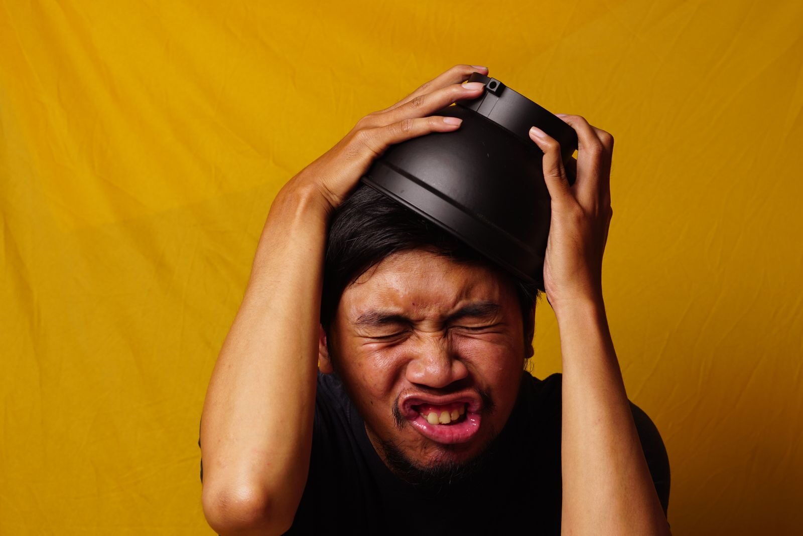 An expressive Indonesian man playfully uses a bowl as a hat against a vibrant yellow background.