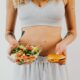 A woman in a sports bra holding a salad and cheeseburger, representing healthy vs junk food choices.