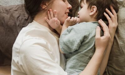 Top view of happy young woman in casual clothes smiling and hugging cute little son while lying together on floor and playing