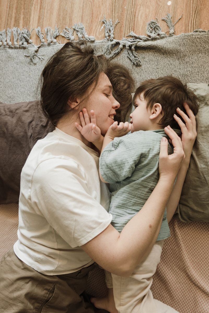 Top view of happy young woman in casual clothes smiling and hugging cute little son while lying together on floor and playing
