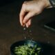 A hand sprinkles salt over a bowl of fresh edamame beans, capturing a moment of delicious meal preparation.