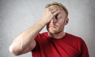 A frustrated man in a red shirt holds his head in stress against a neutral background.