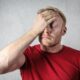 A frustrated man in a red shirt holds his head in stress against a neutral background.