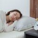 Young calm female with dark hair lying on side with hand under head on white comfortable bed under blanket while sleeping in morning time